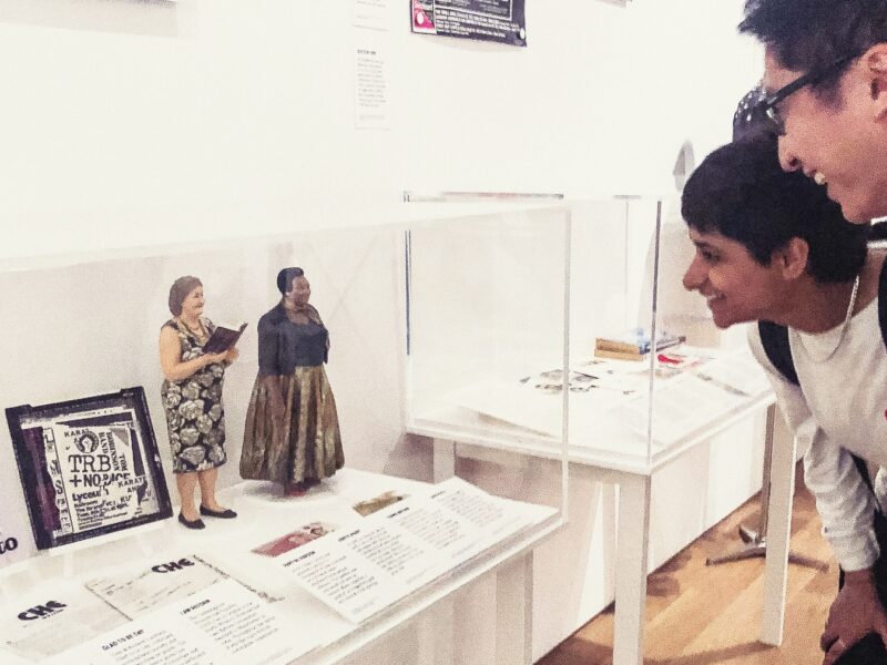 Two museum visitors look intently at a vitrine containing artefacts including two 35cm tall statues of Christine Burns and Lady Phyll