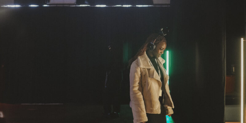 A young woman stands wearing headphones with a movement tracker on top, in front of a green strip light