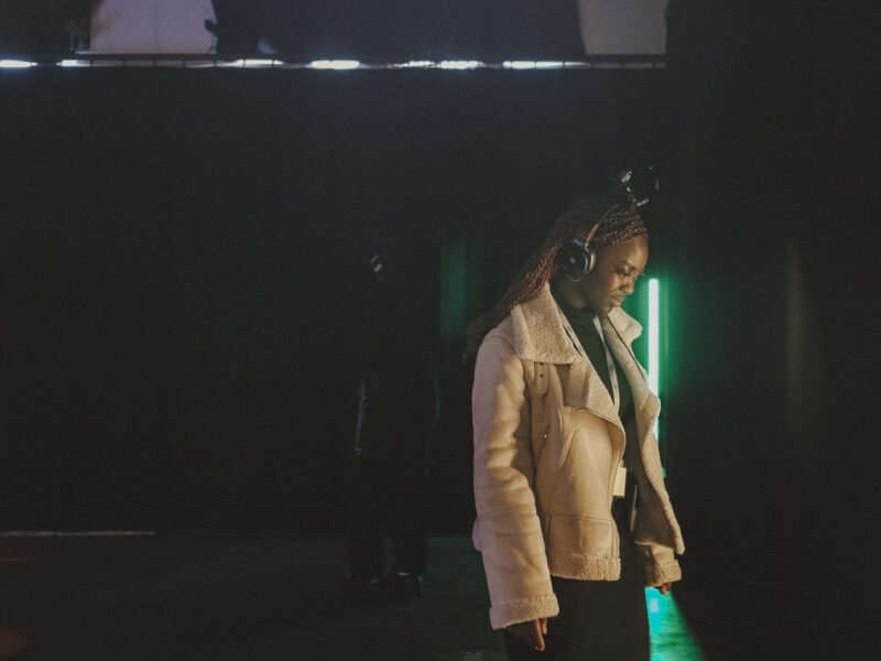 A young woman stands wearing headphones with a movement tracker on top, in front of a green strip light